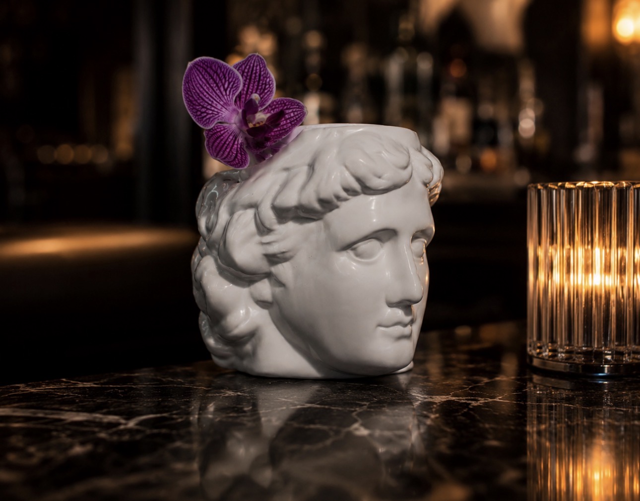 Cocktail served in a white greek goddess head vessel topped with a purple orchid on the marble bar
