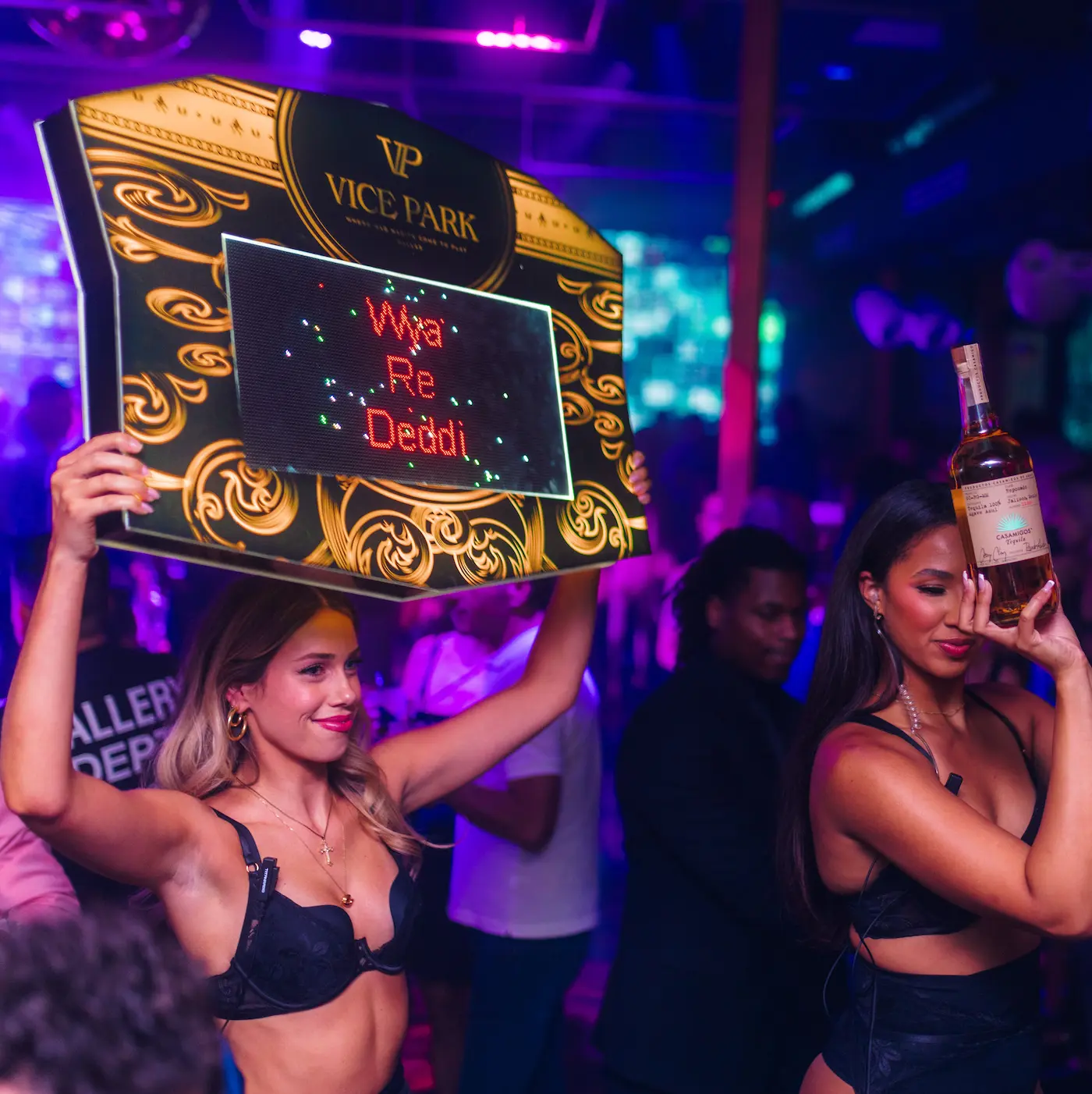 Hostesses presenting a Vice Park LED sign and bottle beneath violet club lighting